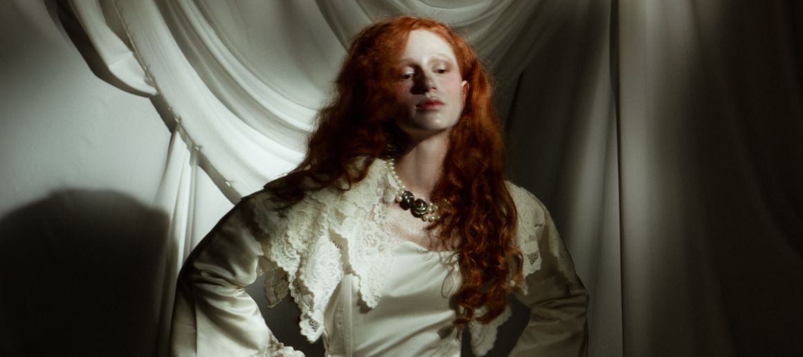Woman in vintage attire with red hair, standing against draped fabric background.