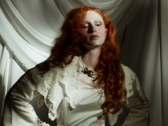 Woman in vintage attire with red hair, standing against draped fabric background.