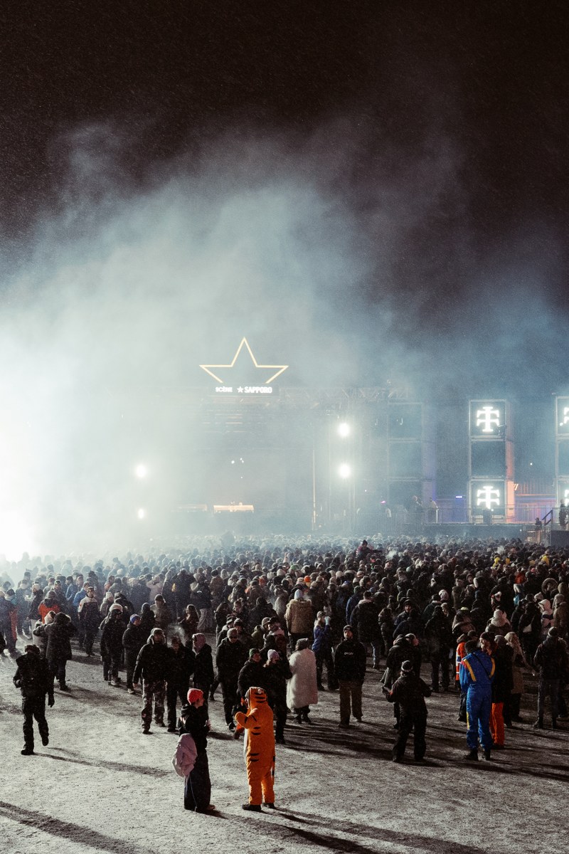Crowd gathers at a foggy outdoor night concert with bright lights in the distance.