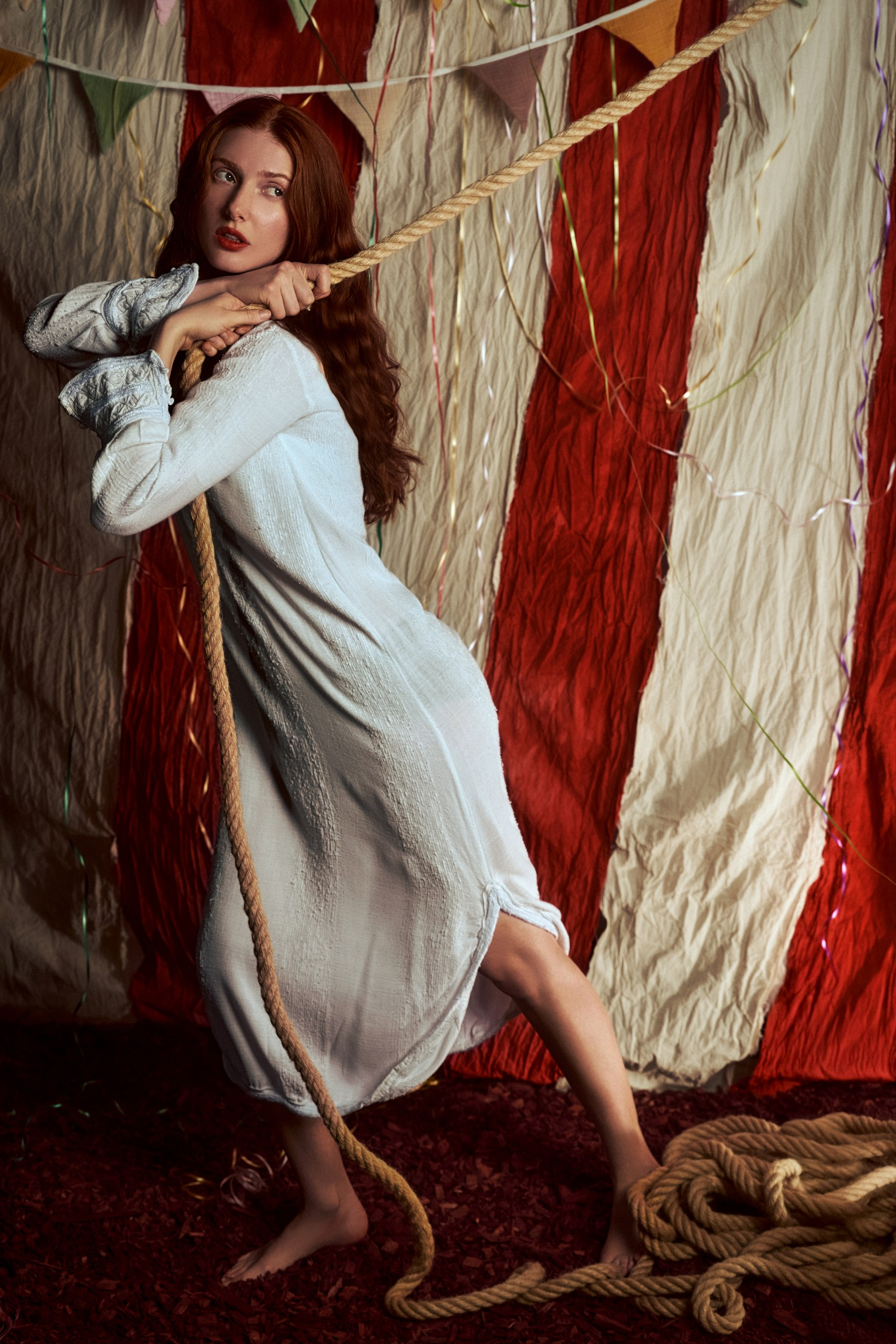 Woman in a white dress pulls rope in a circus-themed setting.