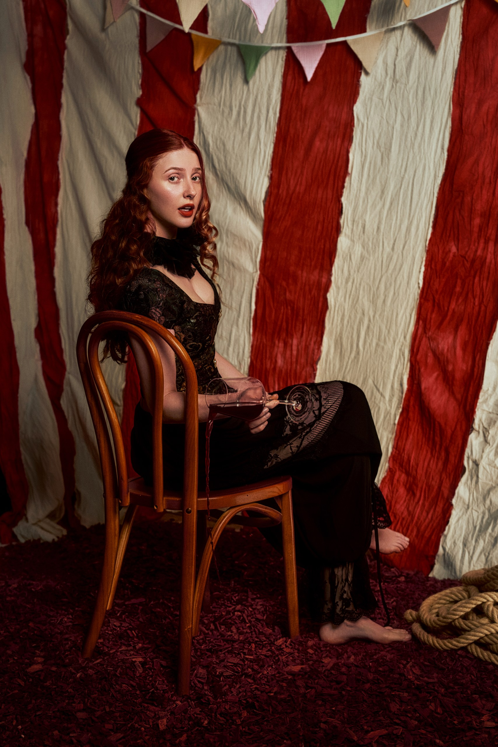 Woman in vintage outfit seated, circus-themed backdrop, spilled wine.