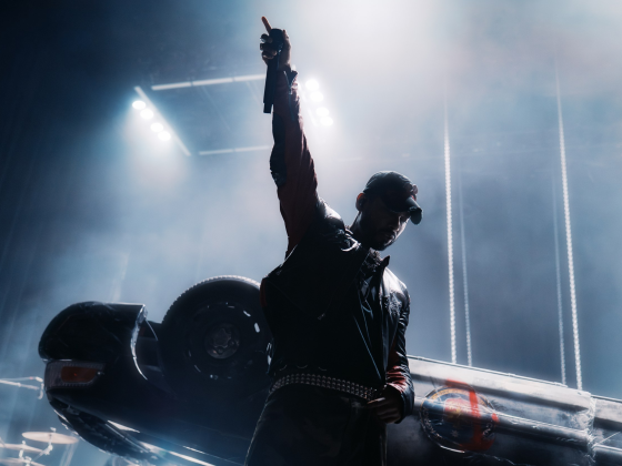 Performer on stage with raised arm, dramatic lighting and overturned car backdrop.