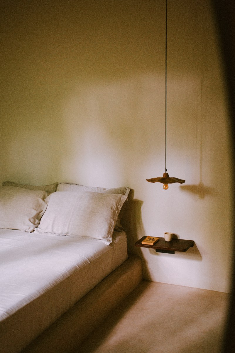 Minimalist bedroom with bed, hanging light, and shelf with book and mug.