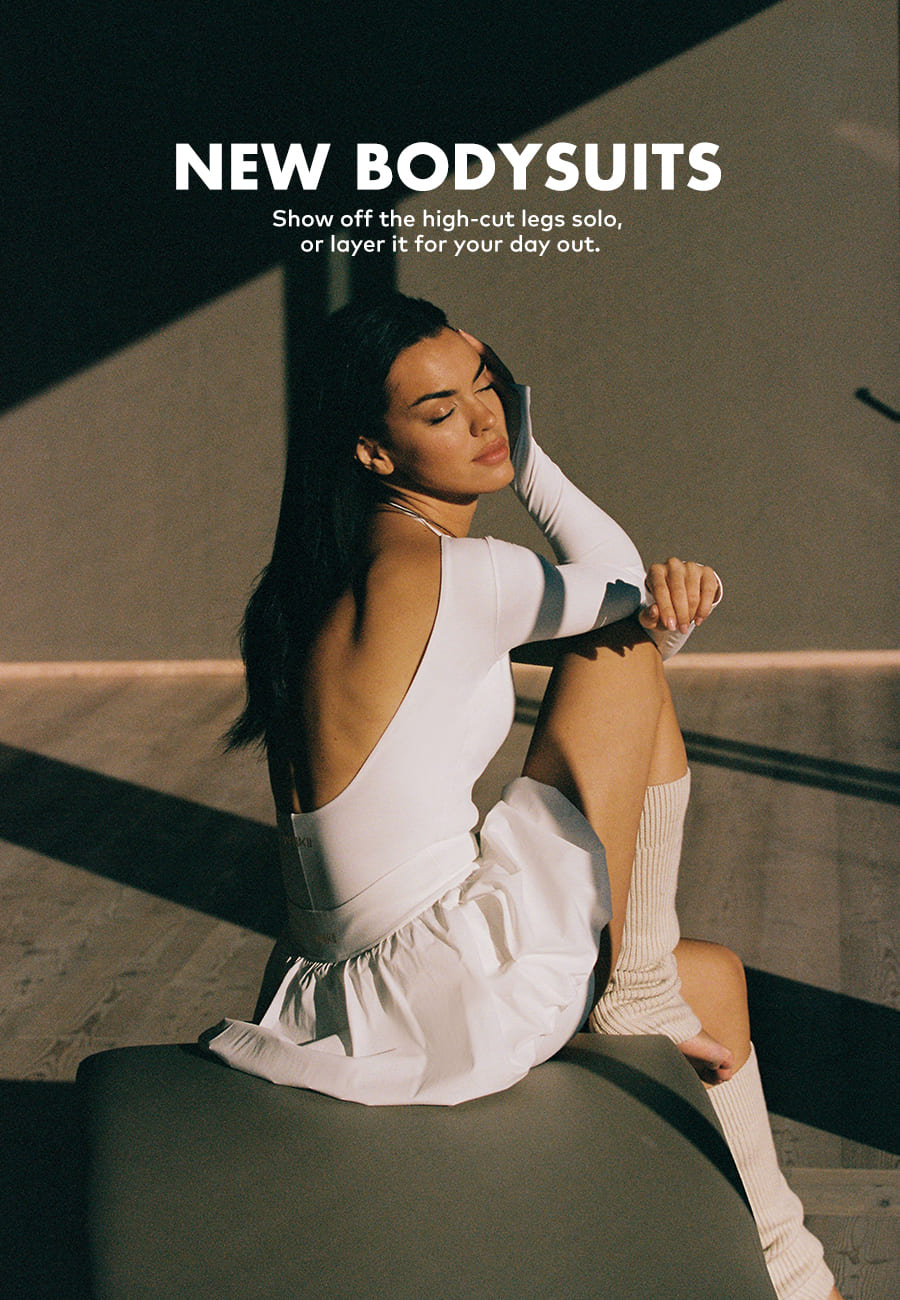 Elegant woman in stylish white bodysuit and socks poses in sunlit room.