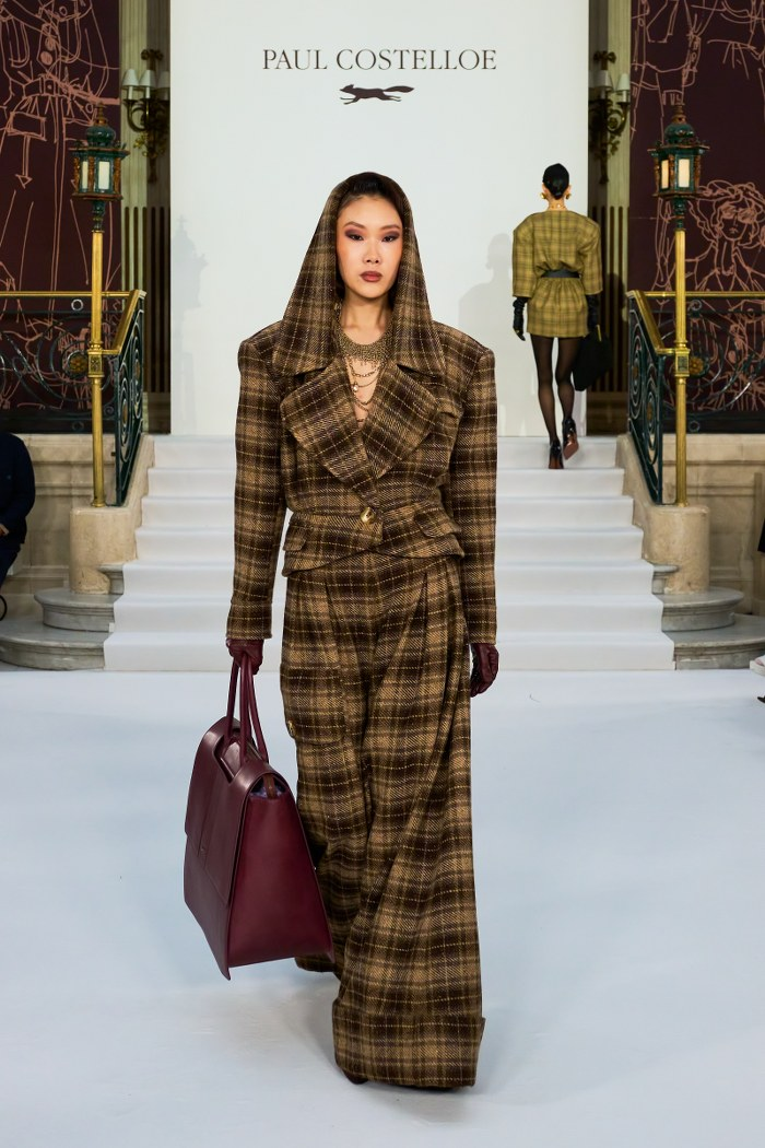Fashion model in hooded plaid coat on runway with burgundy bag.