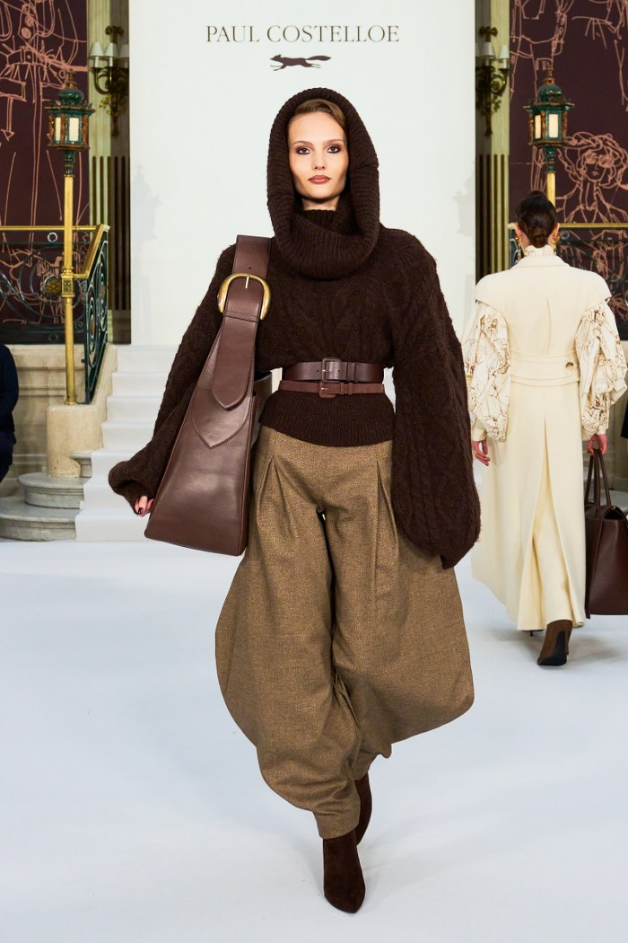 Model on runway in a brown sweater and trousers, carrying a large brown bag.