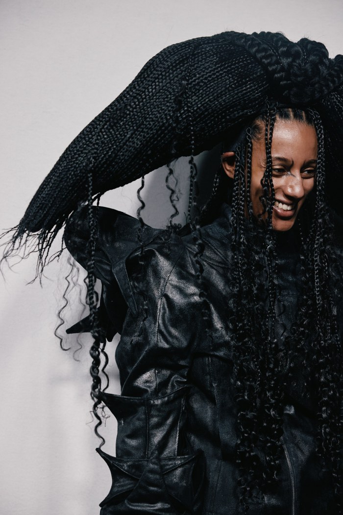 Person with intricate braided hairstyle and black leather outfit smiling.