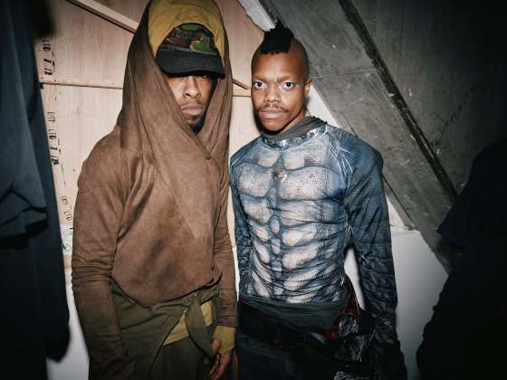 Two men in creative, futuristic outfits under dramatic lighting.