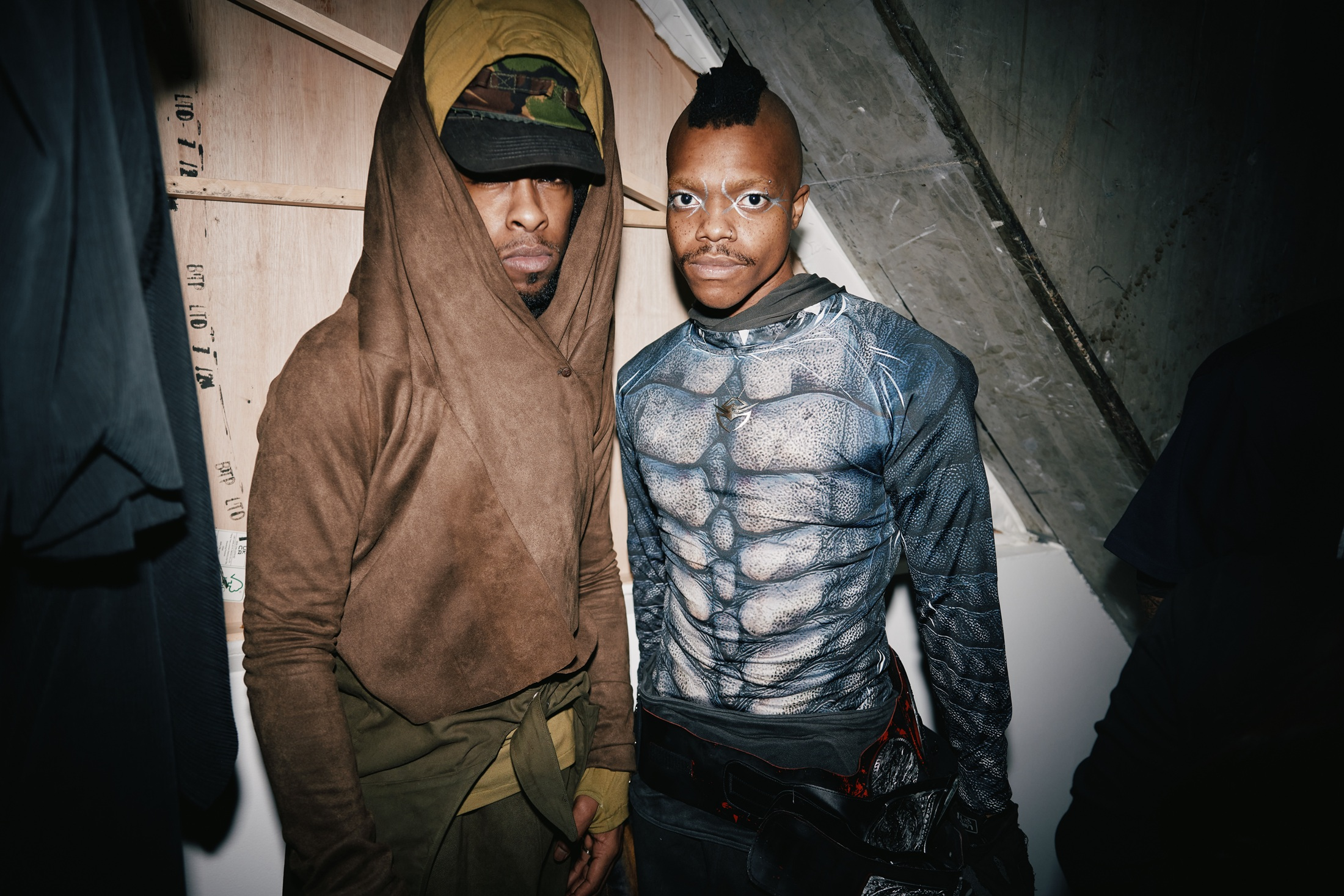 Two men in creative, futuristic outfits under dramatic lighting.