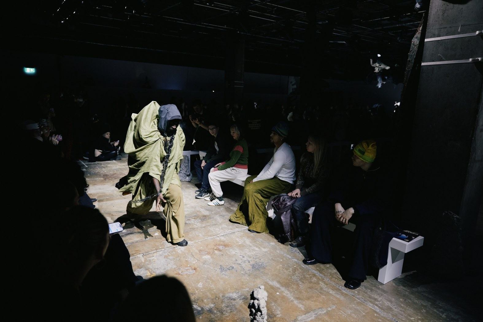 Avant-garde fashion show with unique green-clad model on dimly lit runway.