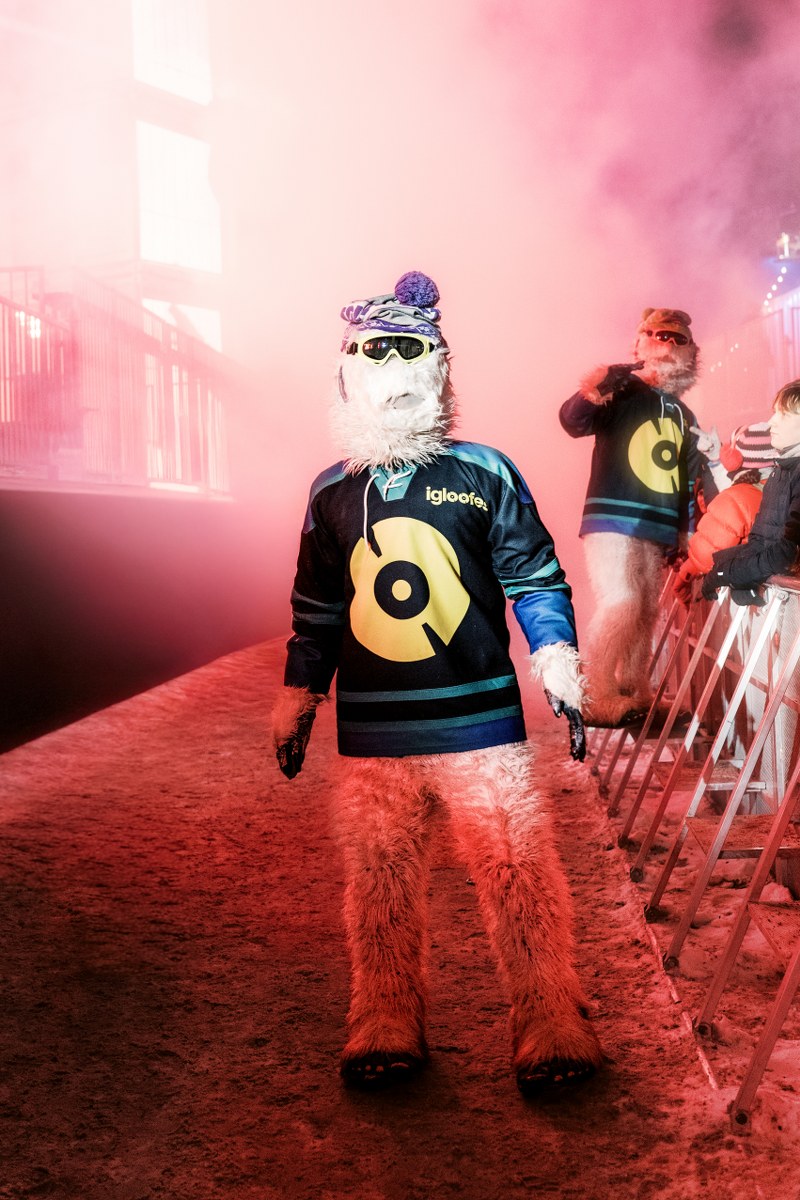 Furry character in winter sports jersey at a snowy, red-lit outdoor event.