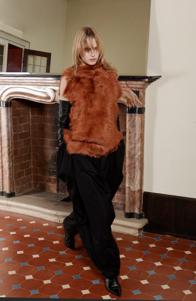 Model in a fur vest and black pants walking indoors by a vintage fireplace.