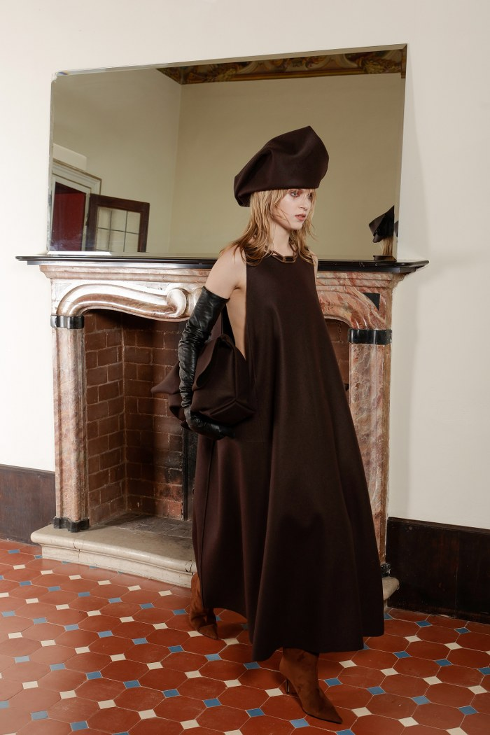 Model in elegant dark brown dress and beret by a vintage fireplace.