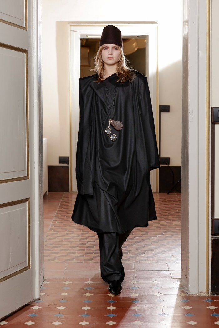 Model in dark avant-garde fashion walking down tiled hallway.