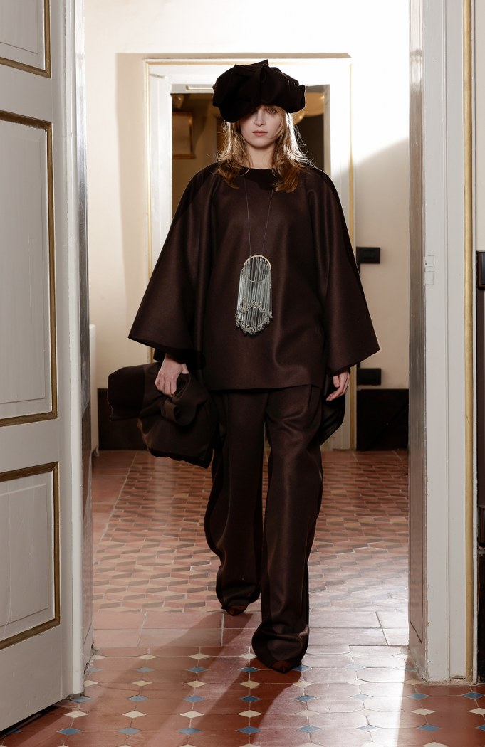 Model in dark brown ensemble with matching hat, walking on tiled floor.