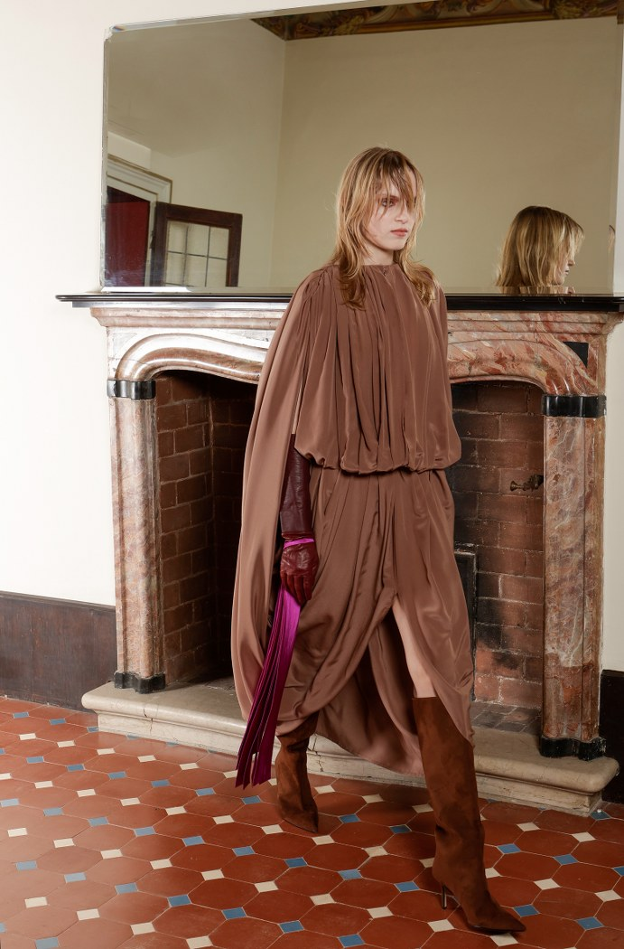 Fashionable woman in brown cape dress, mirror reflection in elegant room.
