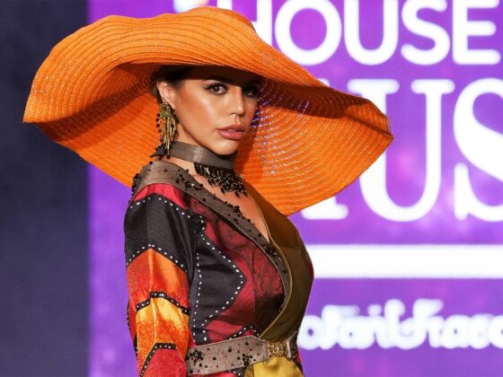 Model in vibrant outfit and orange hat on fashion runway.