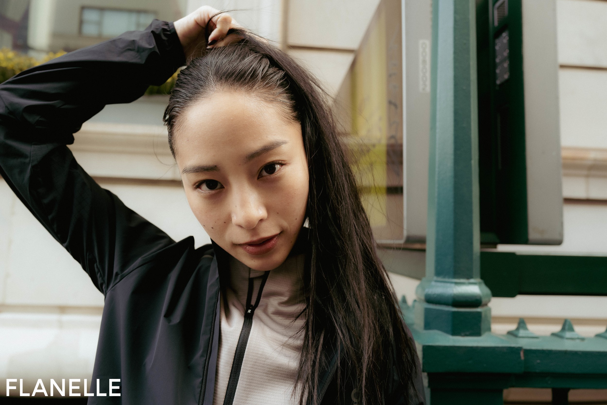 Woman outdoors adjusting hair, wearing a black jacket, urban background.