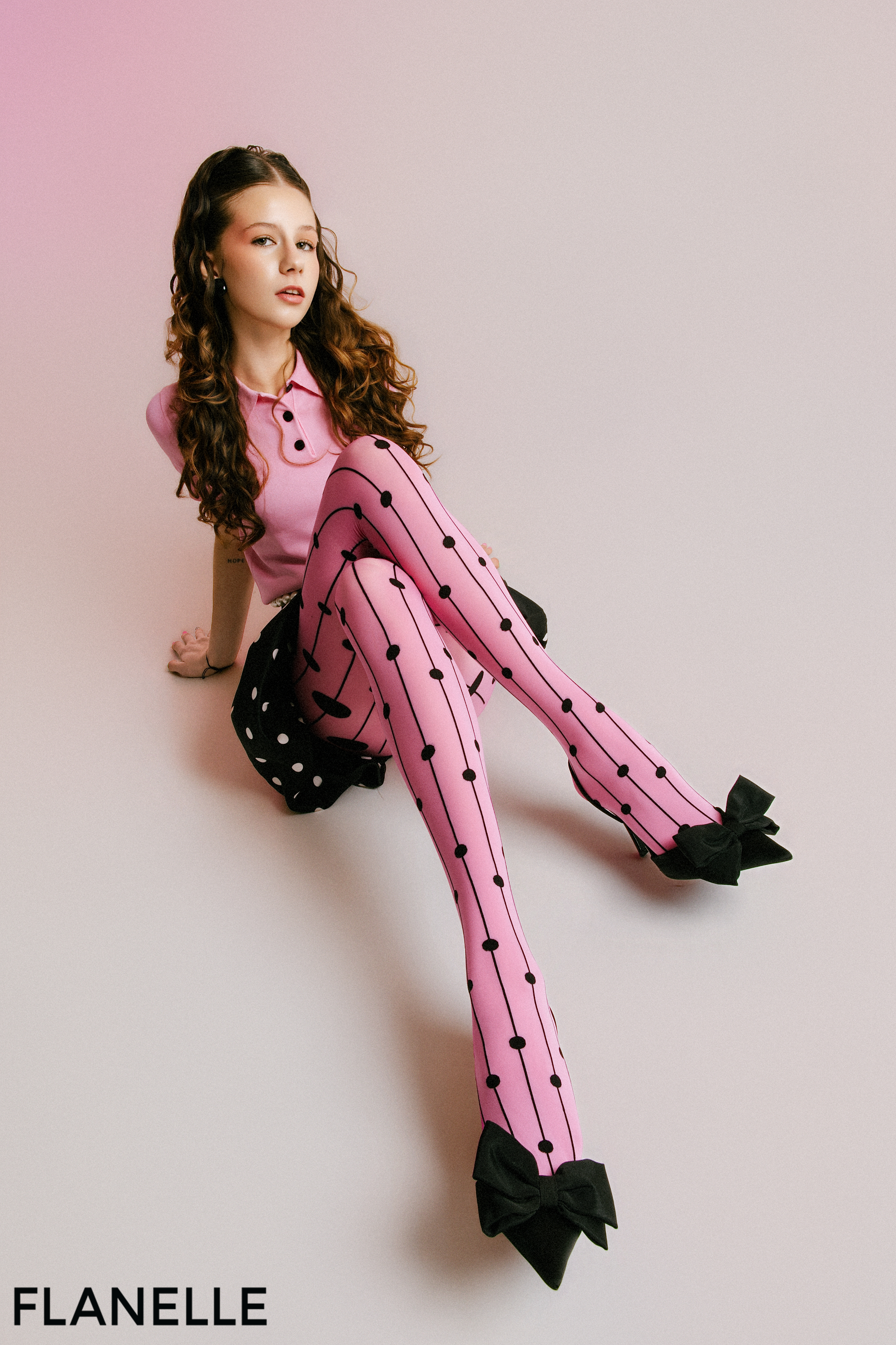 Model in pink outfit with polka dot tights and black bows, seated on pink background.