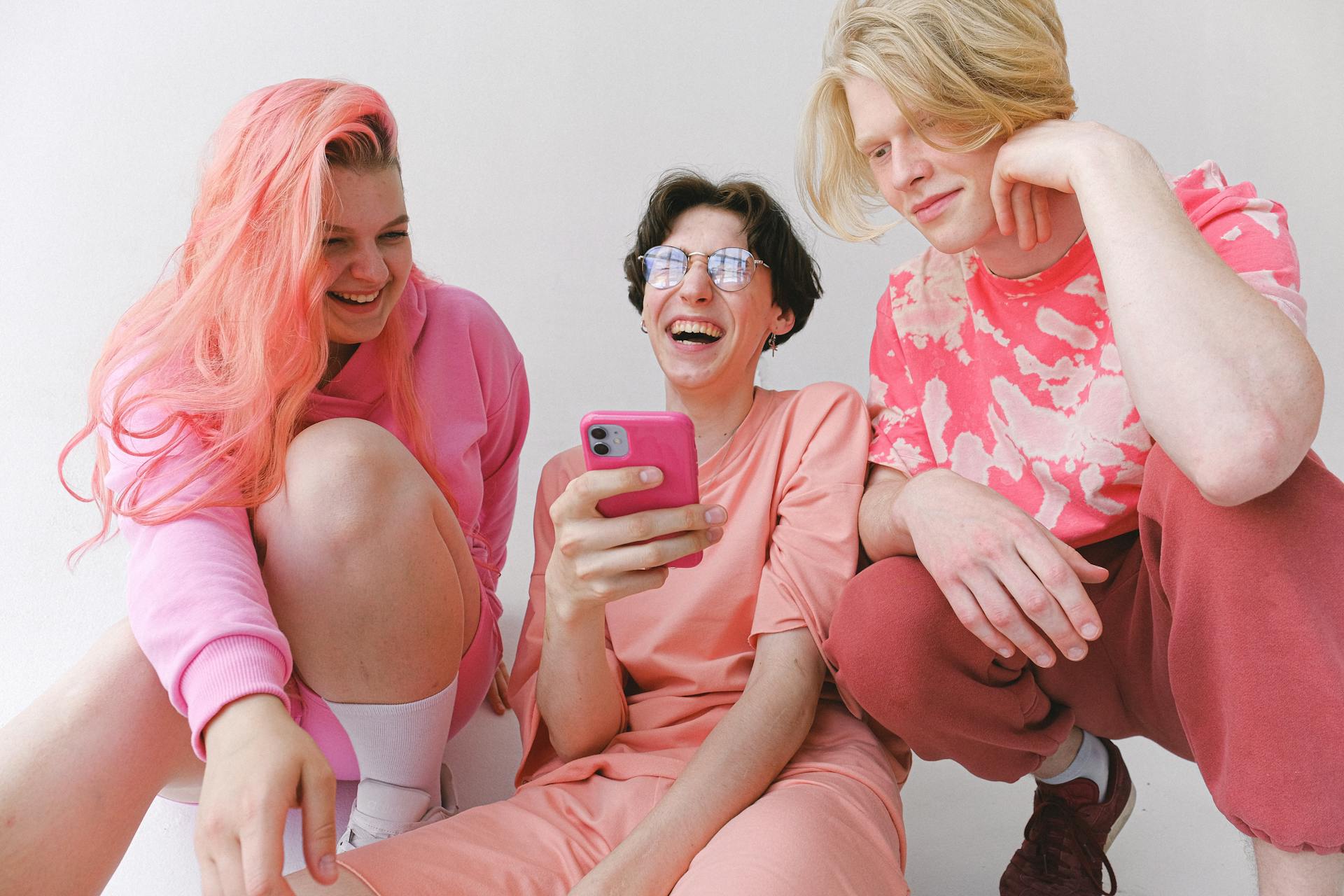 Three friends laughing while looking at a phone, dressed in pink outfits.