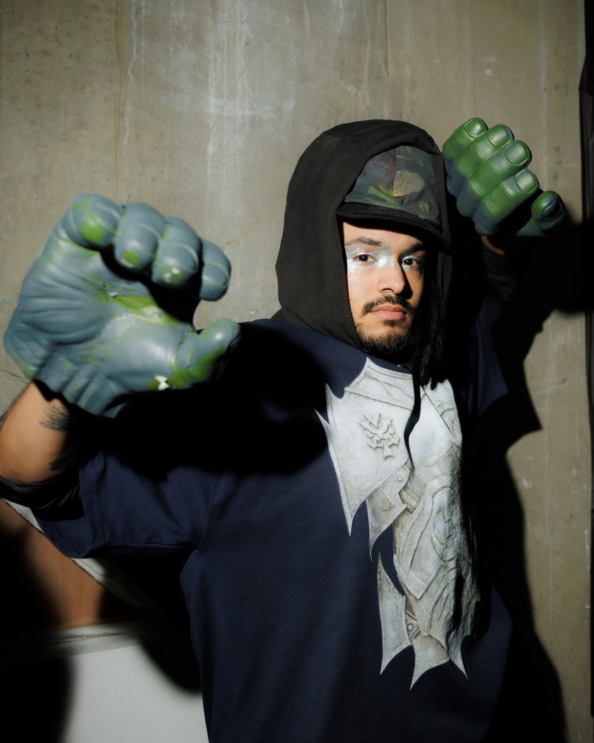 Person in costume with green gloves and hood poses defiantly against a concrete wall.