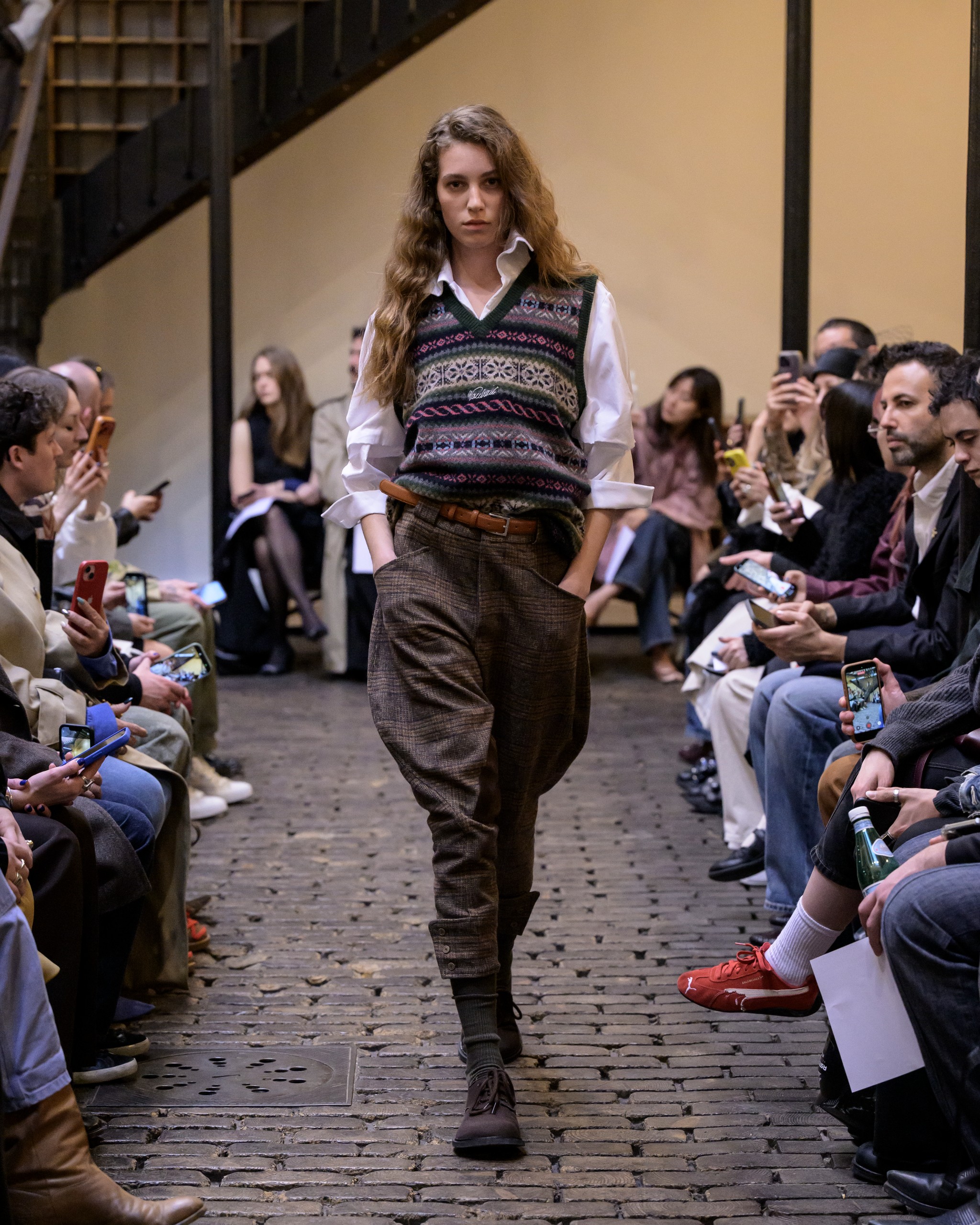 Fashion runway model in layered outfit with plaid pants and patterned vest.