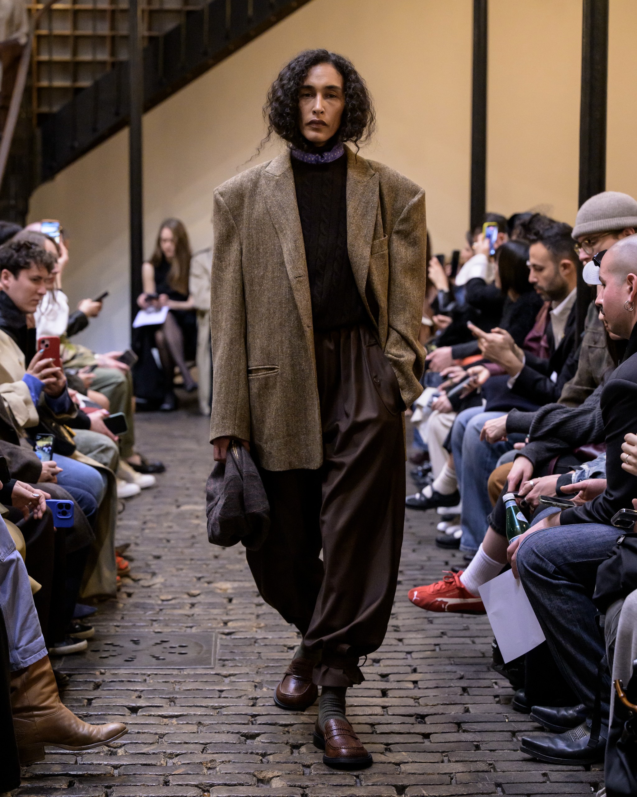 Model walks runway in oversized brown blazer and trousers at fashion event.