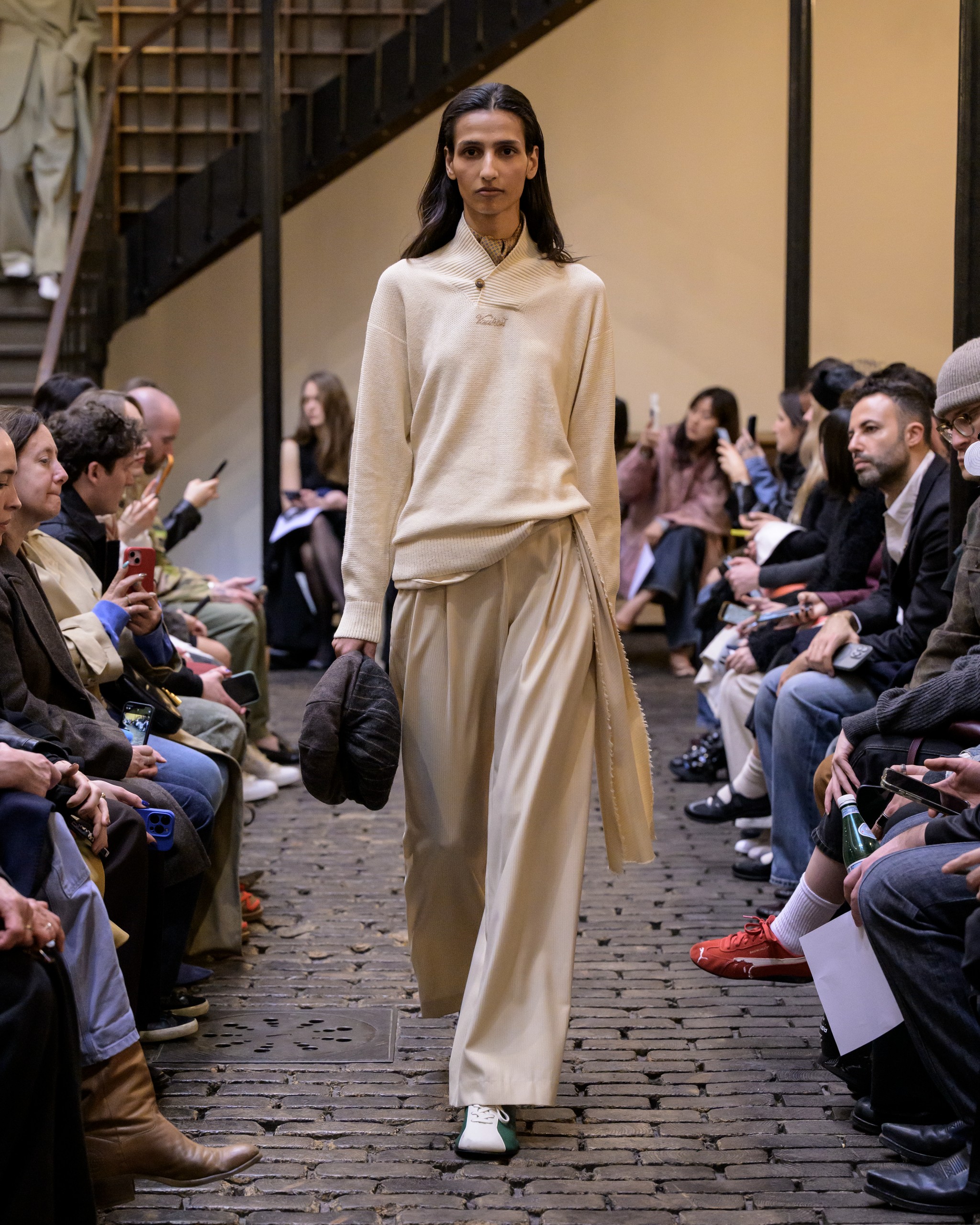 Model in beige outfit on runway, fashion show audience, urban setting.