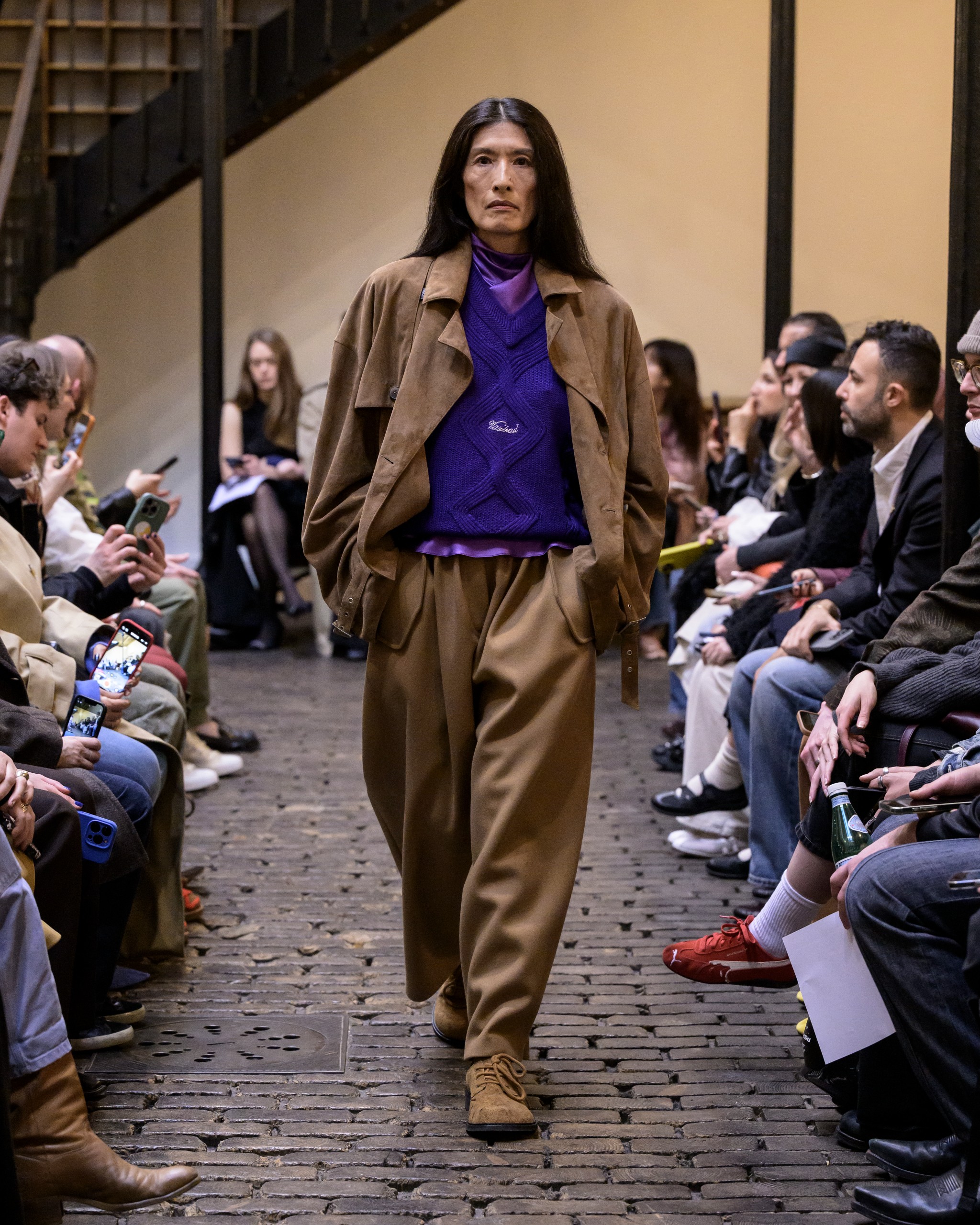 Model in brown outfit walking runway, fashion audience watching.