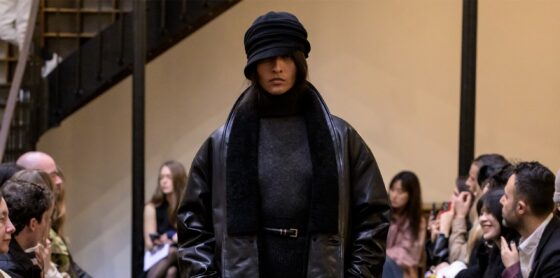 Model showcasing winter fashion at runway event, wearing layered dark attire.