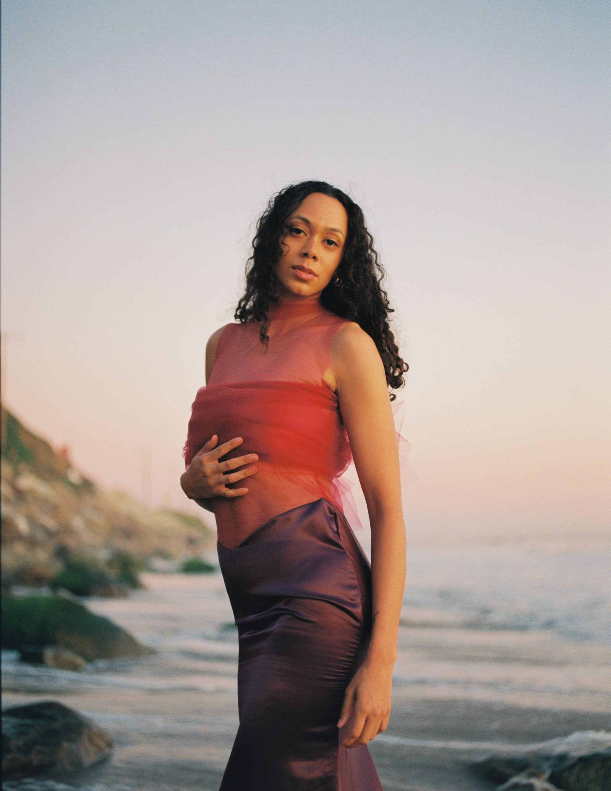 Woman in elegant gown posing by the ocean at sunset.