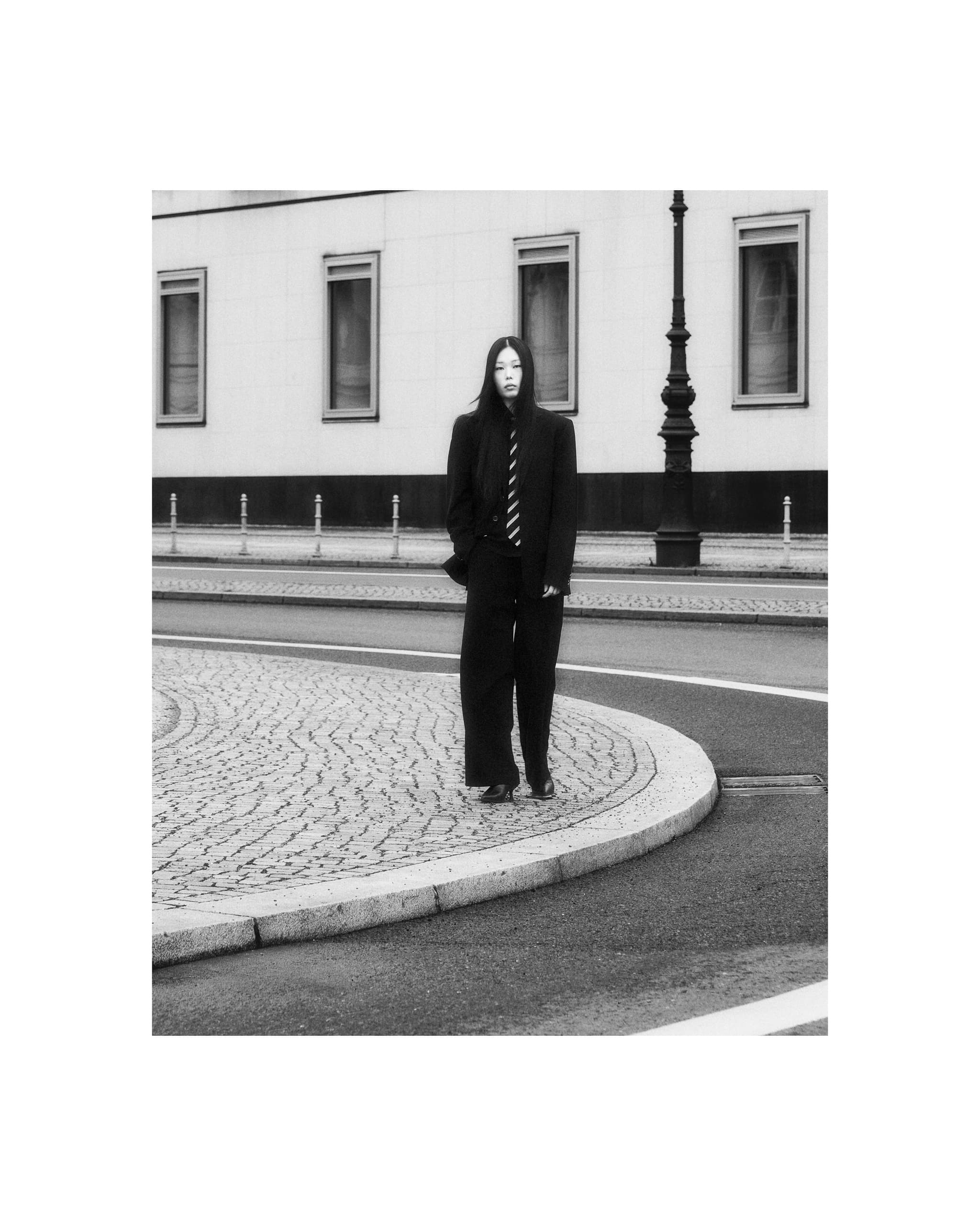 Person in black suit standing on cobblestone street, near building and streetlamp.