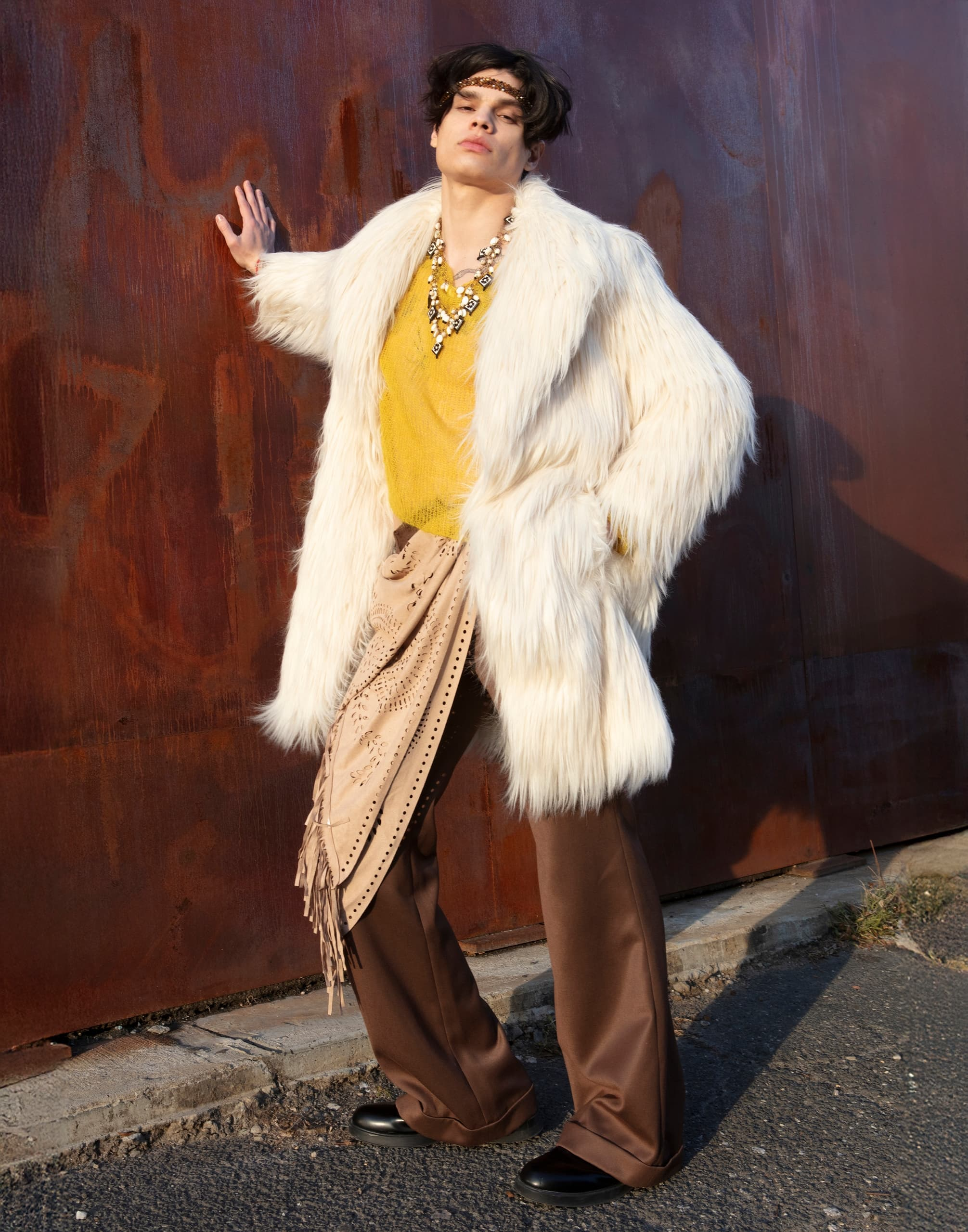 Fashionable person in fur coat and necklace poses against a rustic wall.