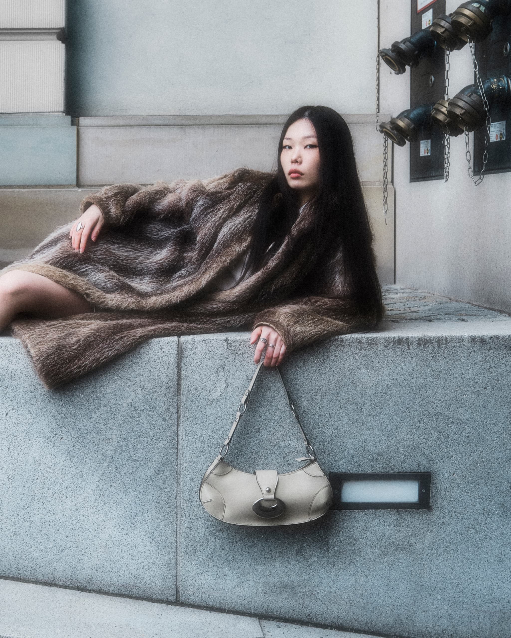 Model in a fur coat lounging with a chic handbag on a concrete surface.