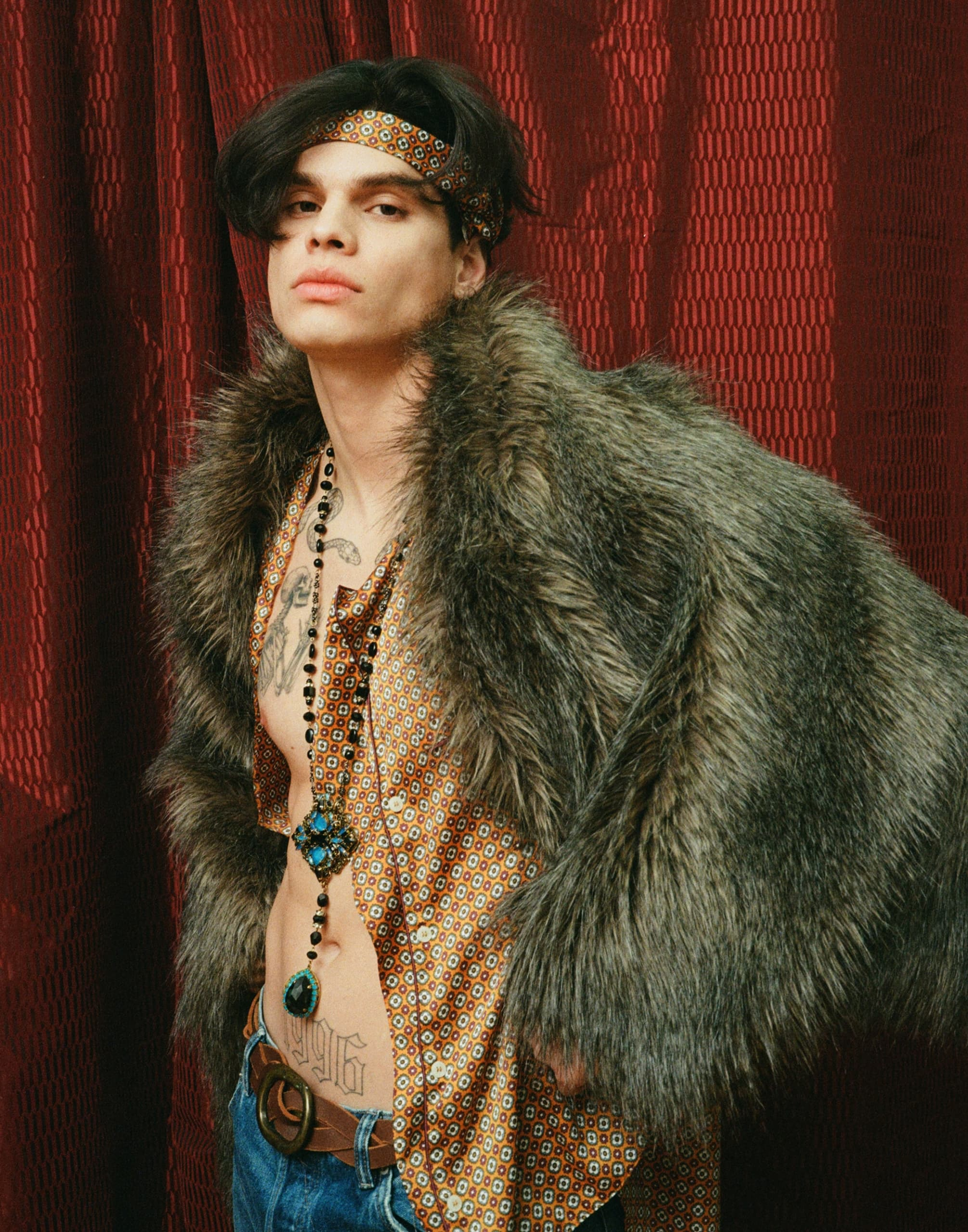 Stylish person in patterned shirt and fur with bold accessories against red curtain.