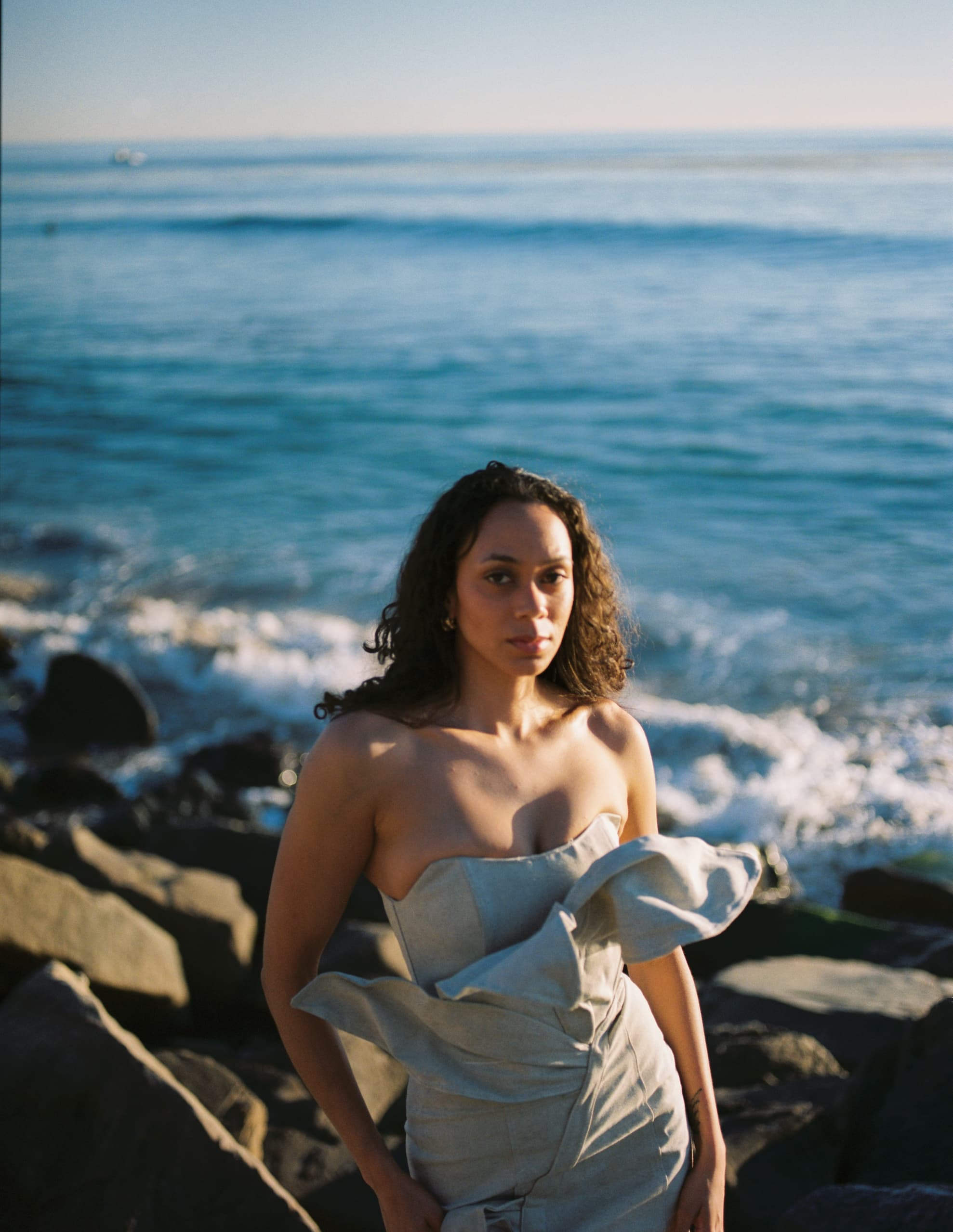 Woman in elegant dress stands by rocky ocean shore under the sun.