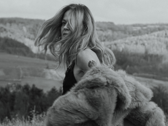 Woman in fur coat with tattoo, windswept hair, stands in open landscape.