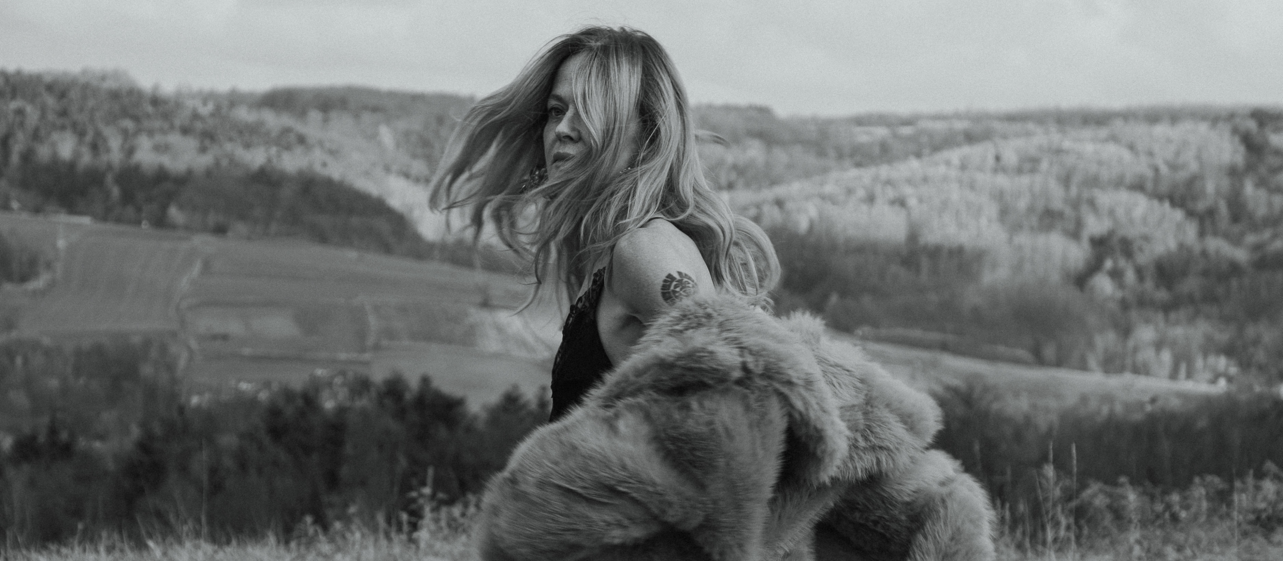 Woman in fur coat with tattoo, windswept hair, stands in open landscape.