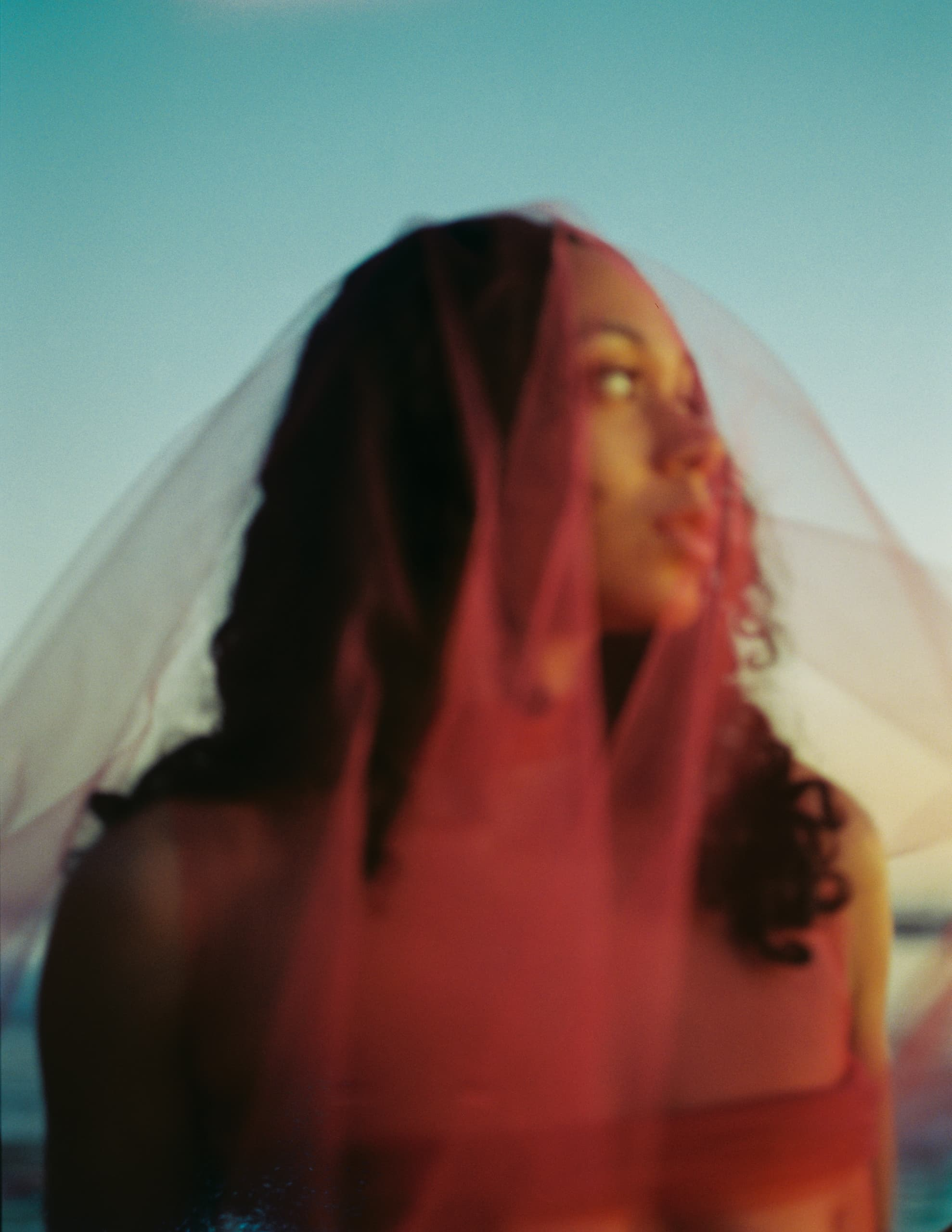 Woman under pink veil gazing into distance against a blue sky.