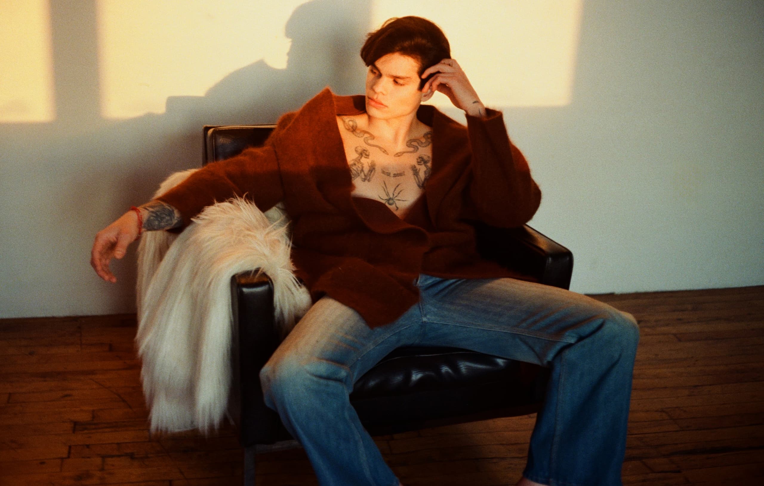 Man with tattoos wearing brown sweater seated in chair, moody sunset lighting.