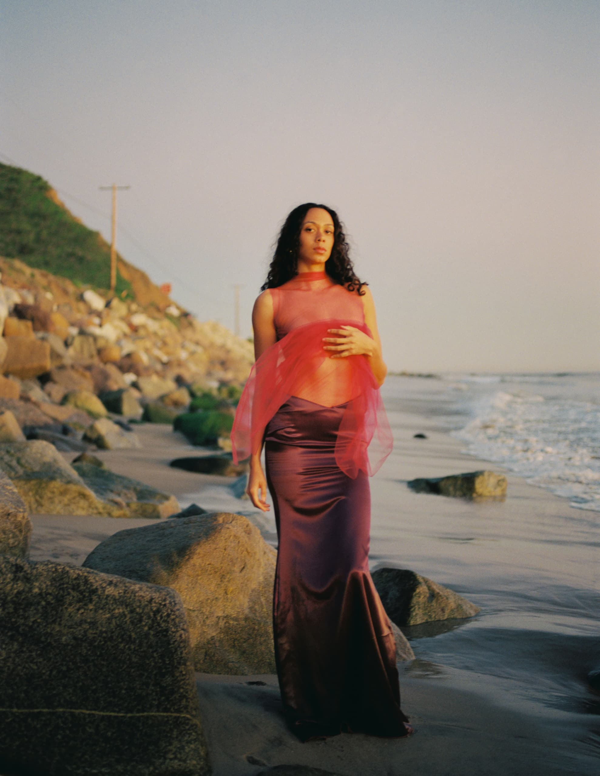 Woman in elegant dress stands on rocky beach at sunset, ocean waves in background.