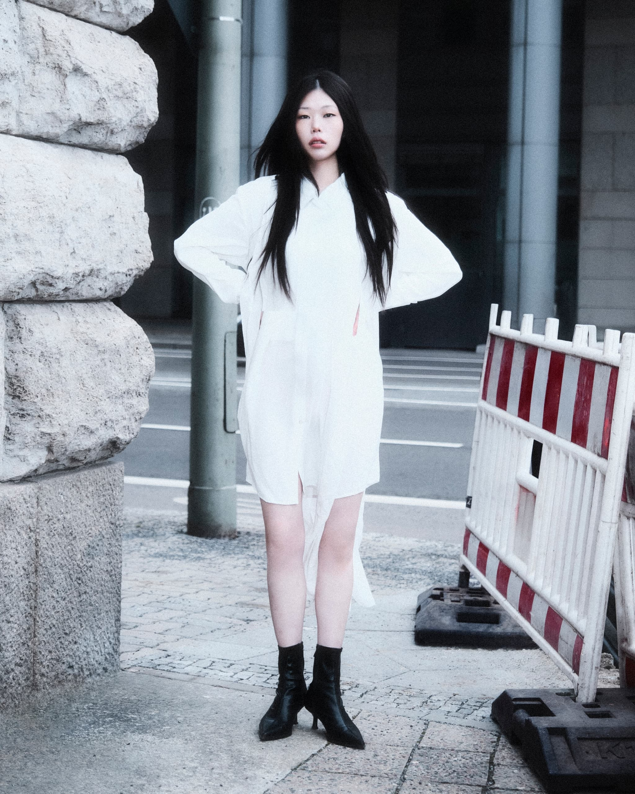 Fashionable woman in white dress and black boots standing on urban sidewalk.
