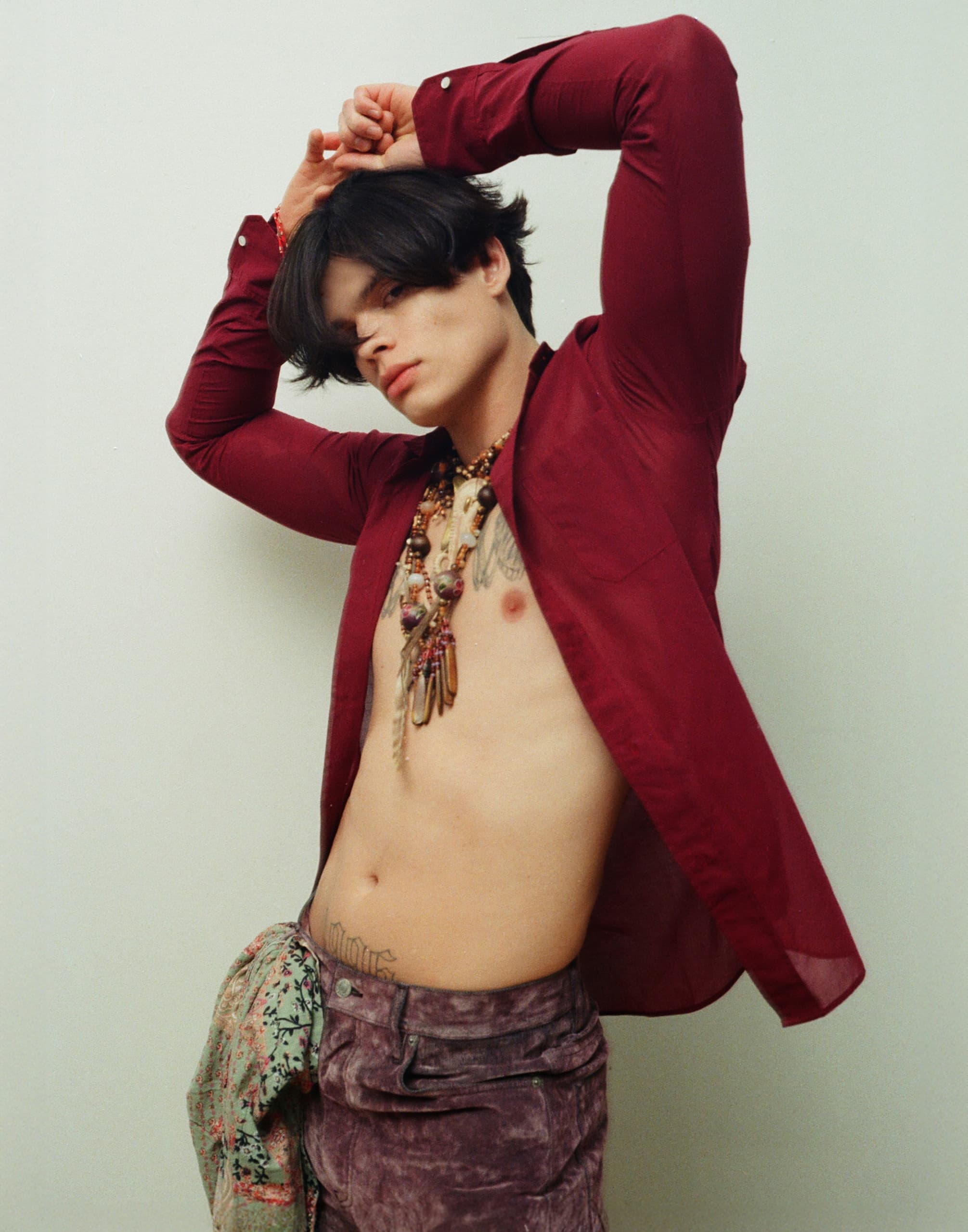 Man poses creatively in open red shirt, wearing layered necklaces. Fashion statement.