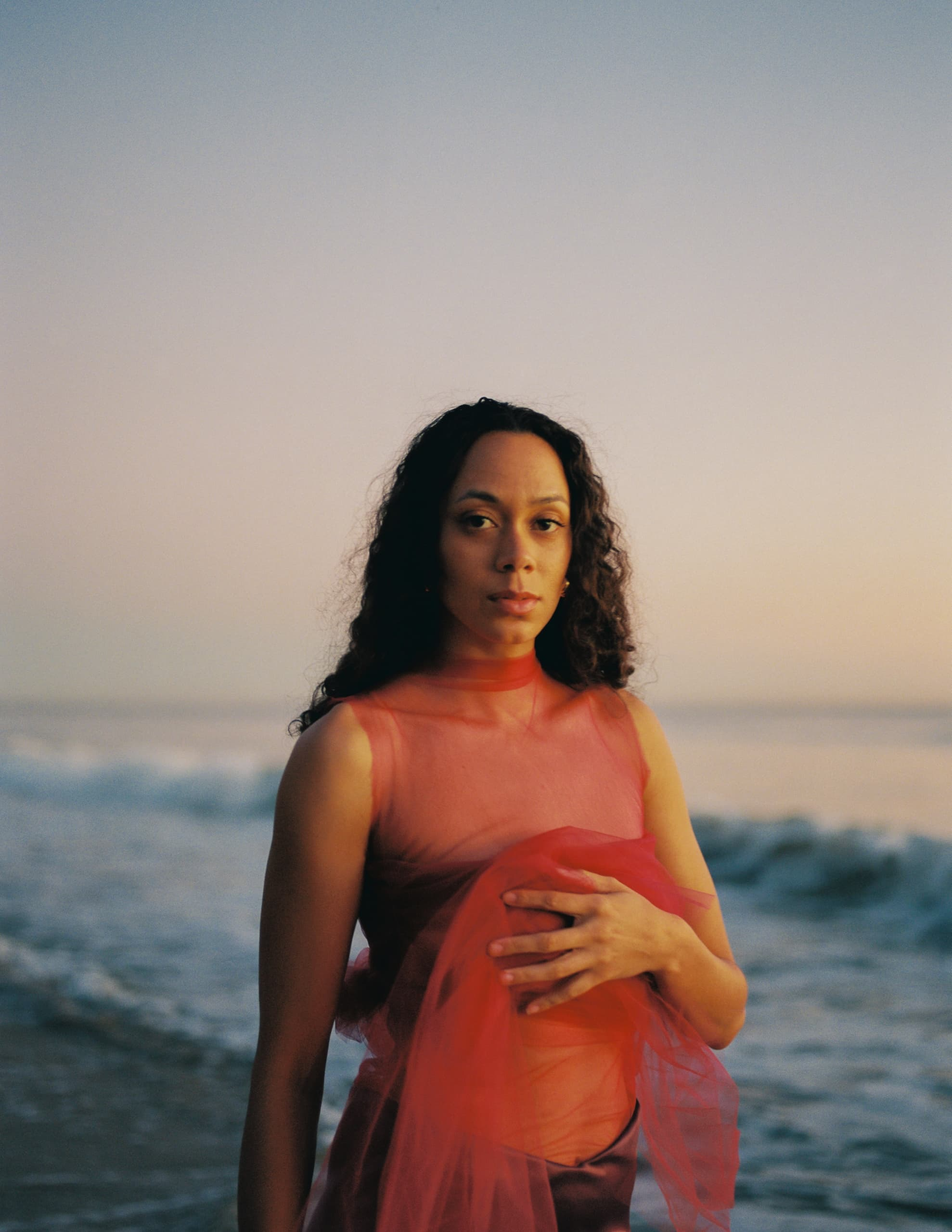Woman in red dress by the ocean at sunset, serene and contemplative.