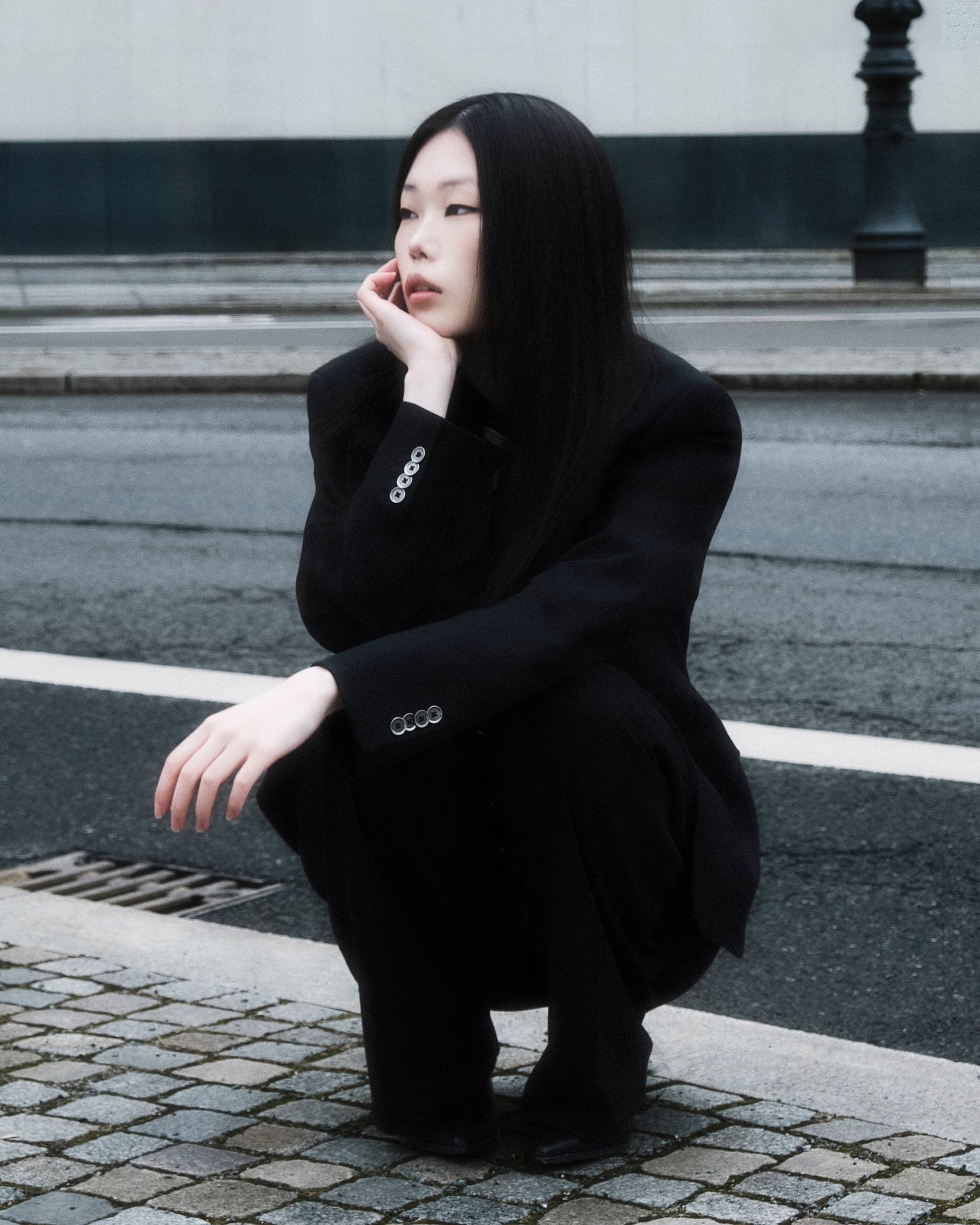 Person in black suit crouching pensively on city street.
