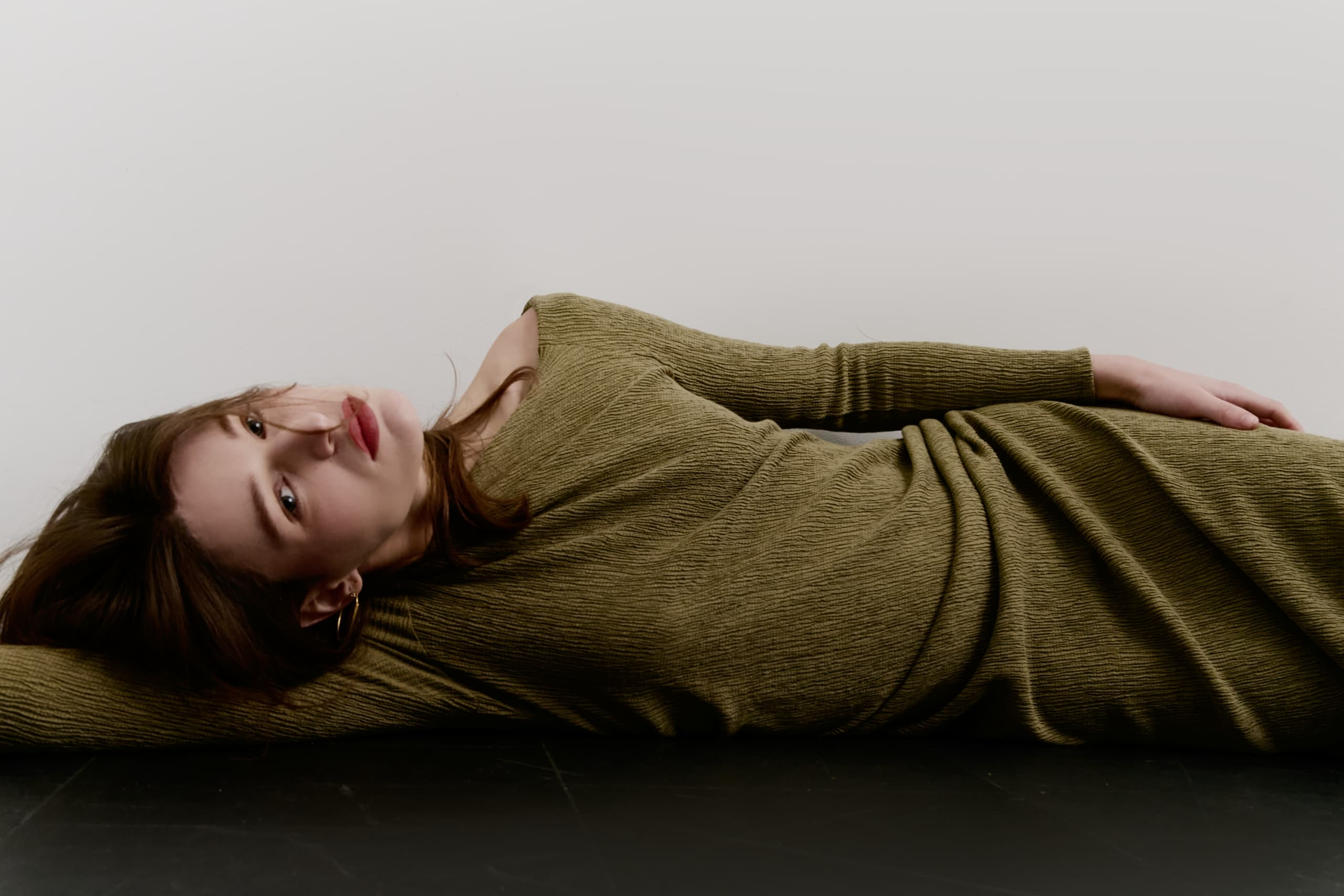 Woman in olive dress lying on the floor, arms extended, looking serene.