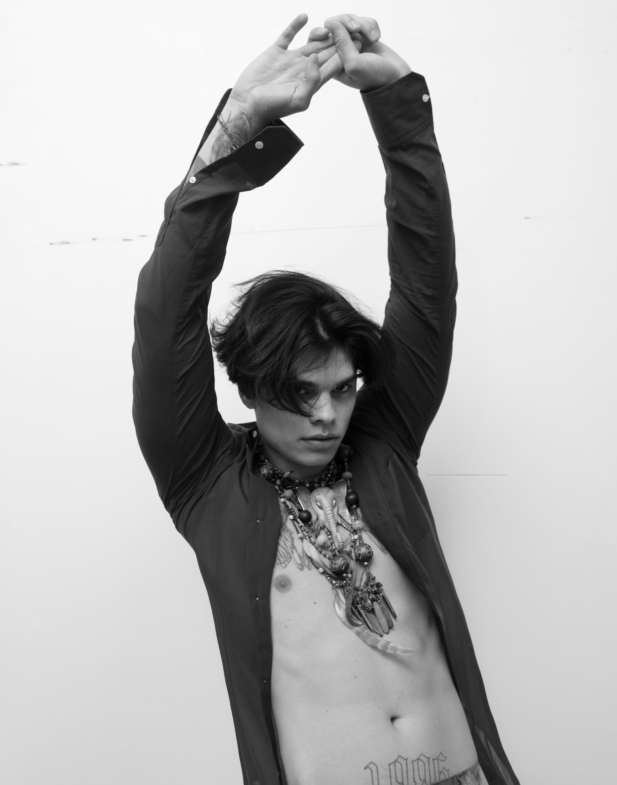 Person posing with hands raised, wearing open shirt and statement necklaces. Stylish portrait.