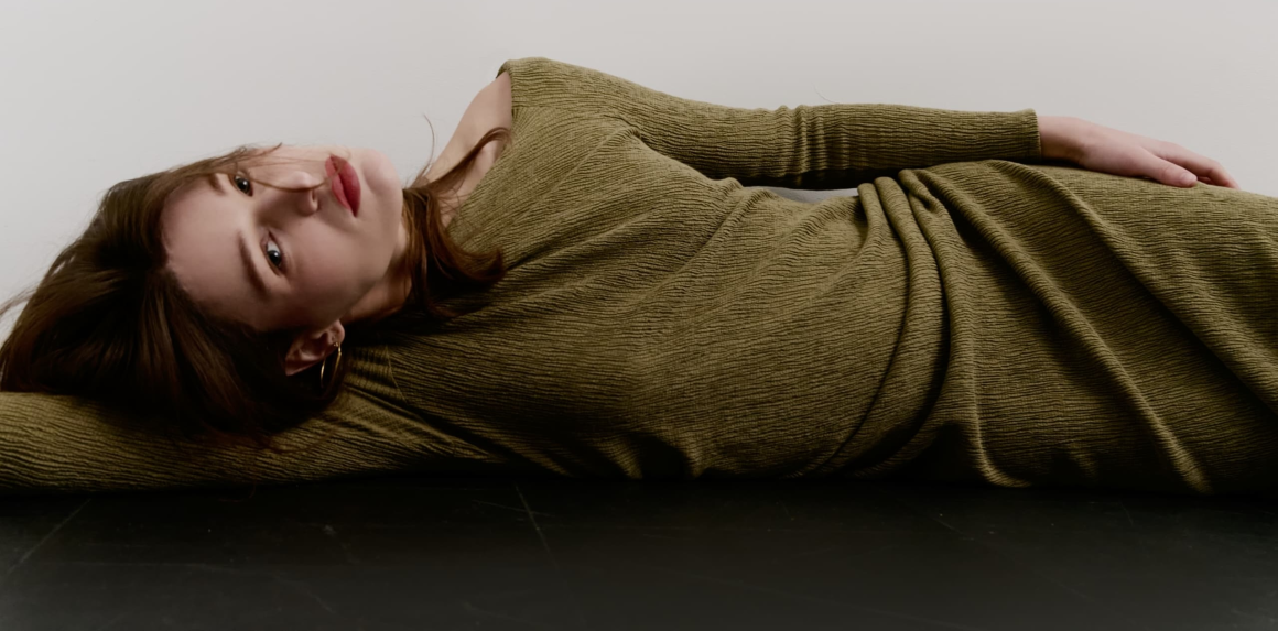 Woman in olive dress lying down on black floor, looking contemplative.