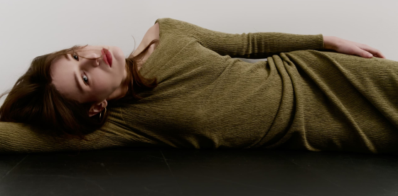 Woman in olive dress lying down on black floor, looking contemplative.
