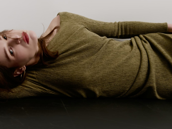 Woman in olive dress lying down on black floor, looking contemplative.